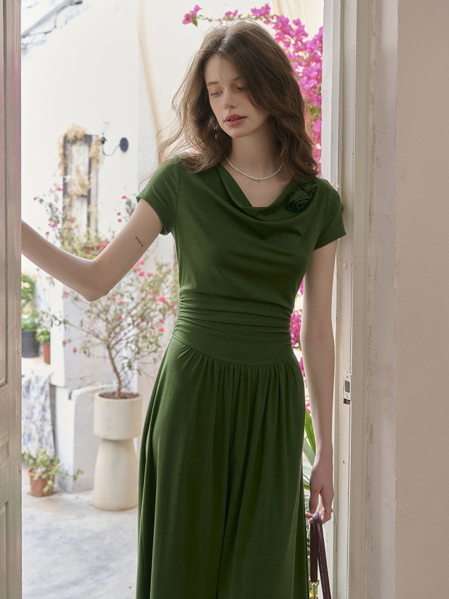 Model in a soft green draped-neck dress with a cinched waist and flowing skirt, holding a small burgundy bag while standing by an open door with blooming flowers in the background. Graceful and romantic garden look.