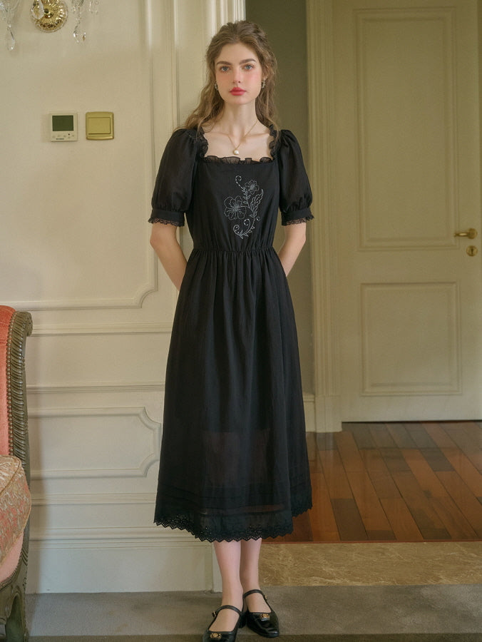A young woman with soft curls and a red headband wearing a black retro vintage dress with floral embroidery, standing gracefully in an elegant room with classic interior details.