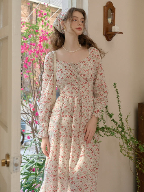 A woman in a vintage floral long dress with a shirred waist and delicate red flower print, styled with a cream net bag, creating a cottagecore retro aesthetic.