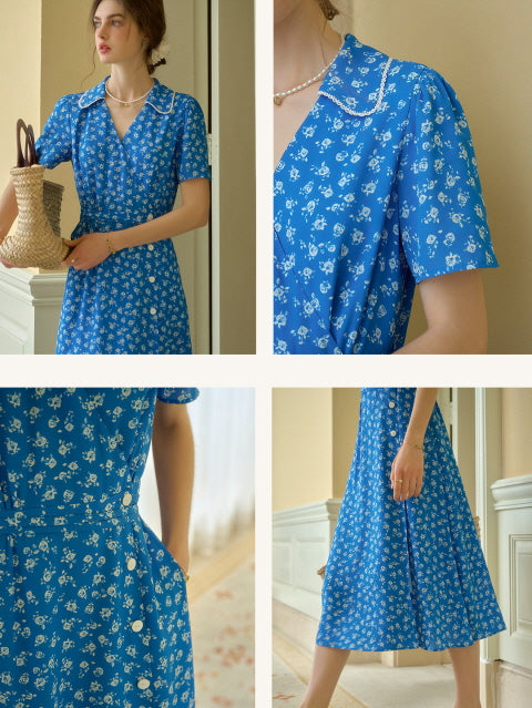 Woman wearing a vibrant royal blue floral midi dress with pearl-trimmed collar and short sleeves, standing indoors in soft natural light with a woven basket bag. Romantic and vintage-inspired summer outfit.