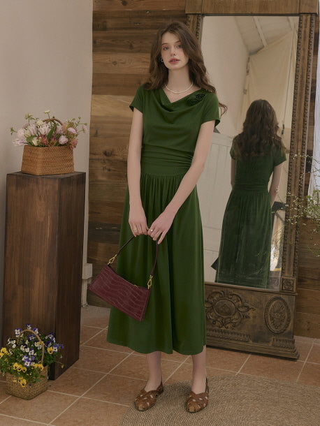 Woman wearing an elegant forest green midi dress with a draped neckline and fitted waist, standing indoors next to a mirror and floral decorations. Romantic vintage-inspired outfit with a serene mood.