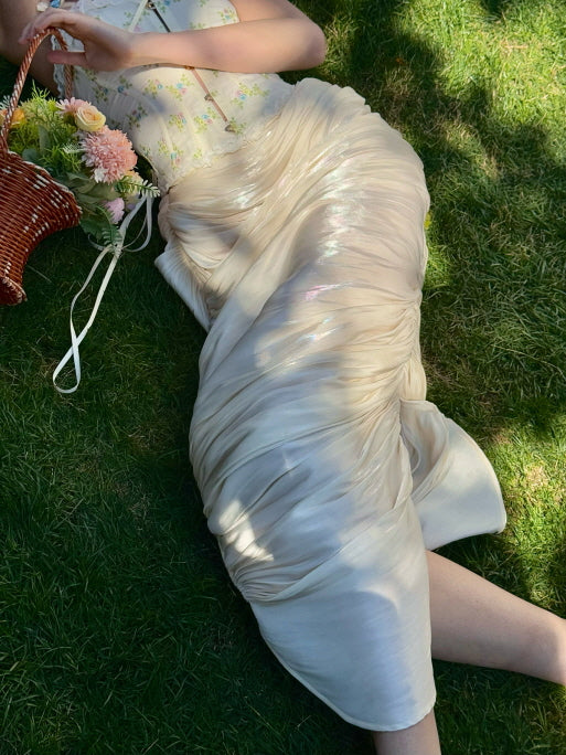 Cream ruched satin midi skirt with glossy finish, styled for a romantic spring picnic look, lying on grass with a wicker basket of pastel flowers – fairytale aesthetic women’s fashion