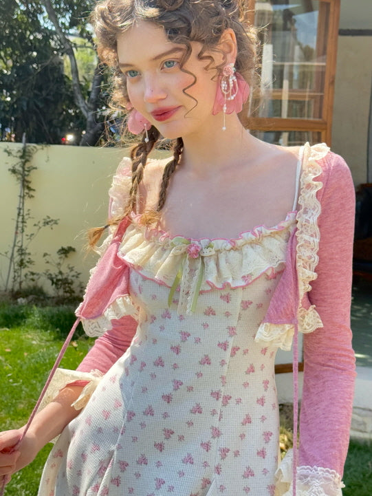 A close-up of a young woman wearing a pink floral-patterned knit dress with lace ruffle detailing, paired with a cropped pink cardigan adorned with lace trim and ribbon ties.