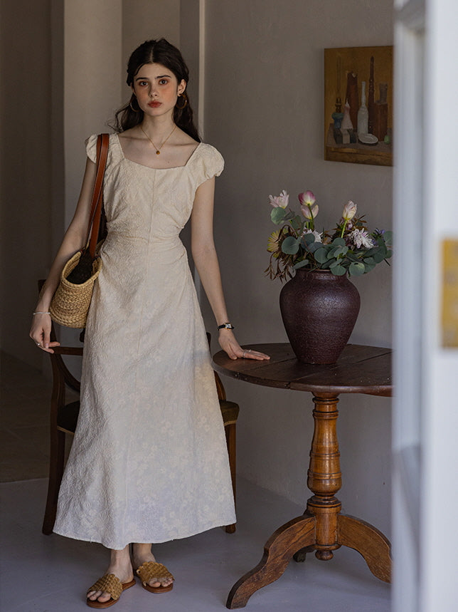 Woman in a cream-colored floral textured midi dress with puff sleeves and a square neckline, carrying a woven tote bag while standing near a wooden table with a vase of flowers in a cottagecore-themed room.