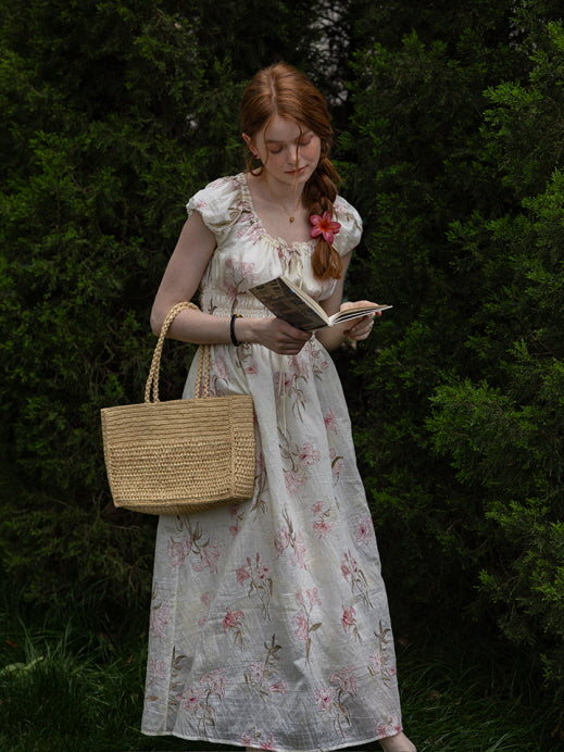 Woman holding a book and woven bag, wearing a floral cottagecore dress with ruffled neckline and flowing silhouette. Dreamy, vintage-inspired look.