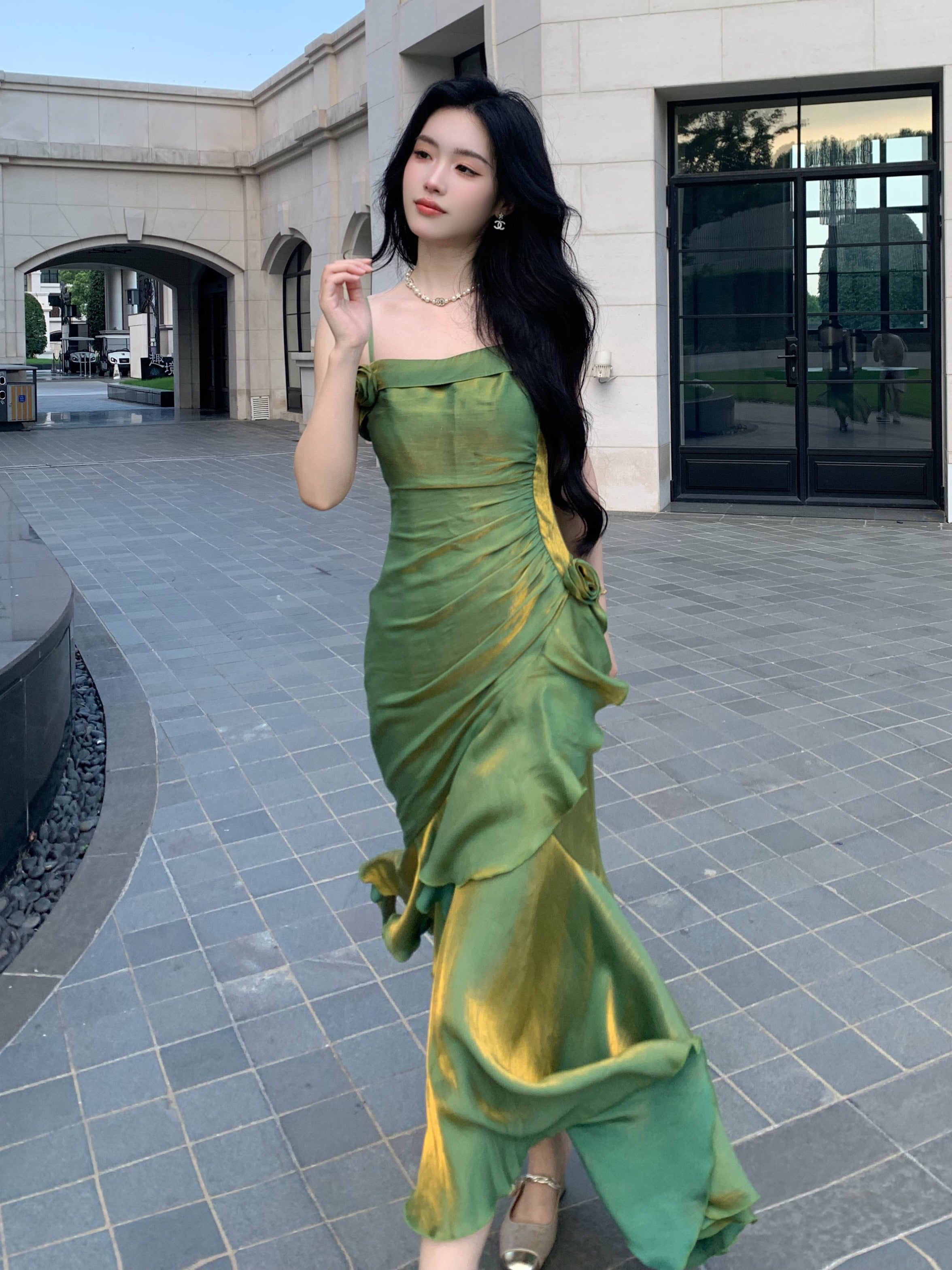 Layered green satin fairytale dress with asymmetric hem and rosette trim, styled in an outdoor walkway.