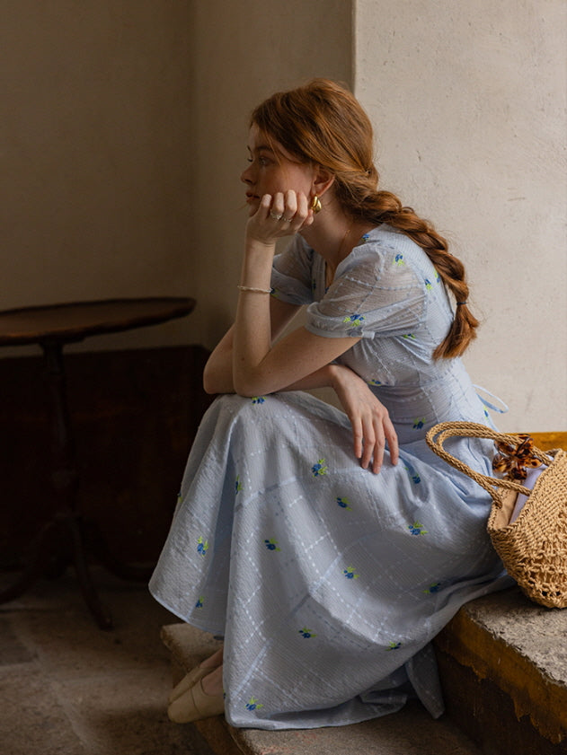 A woman sitting thoughtfully on a stone step, wearing a light blue floral print midi dress with puff sleeves and a delicate check pattern.