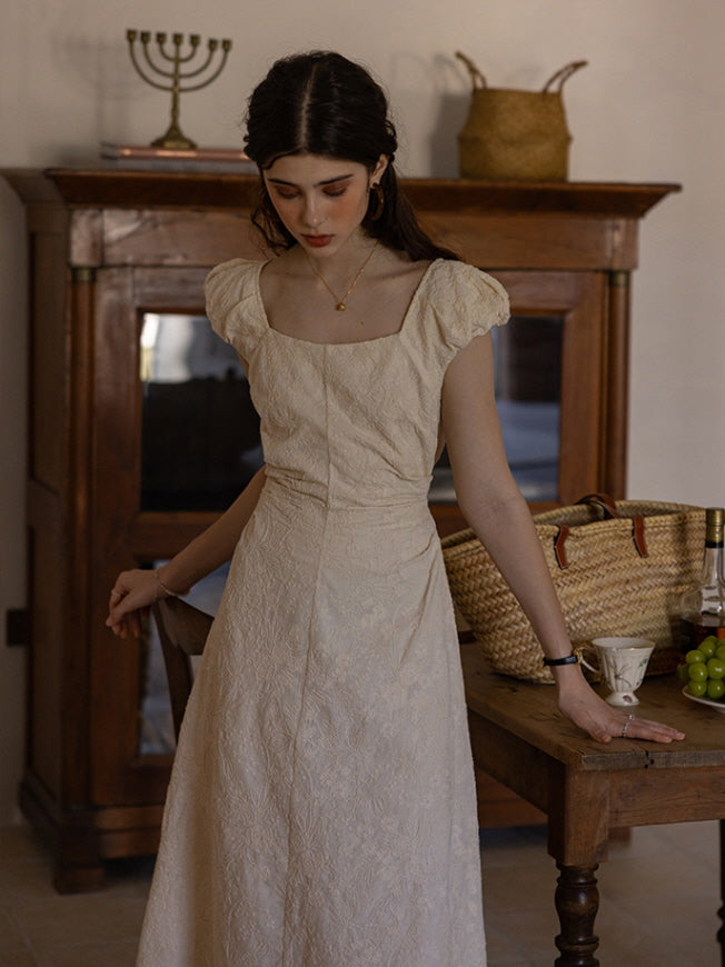 Young woman wearing a cream-colored textured midi dress with puff sleeves and a fitted waist, standing beside a wooden table with a wicker basket and a teacup in a vintage-inspired interior.