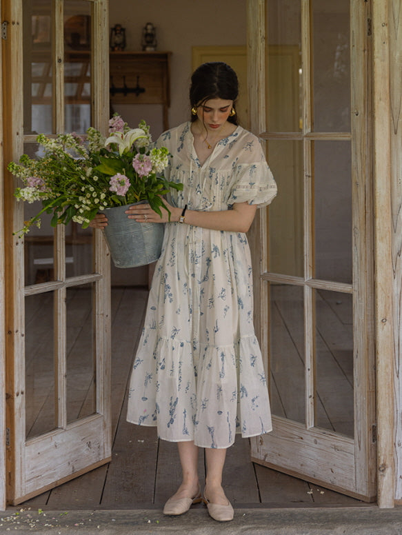 Woman standing in a rustic wooden doorway wearing a cream floral print dress with blue botanical motifs and tiered skirt, holding a large bucket filled with fresh flowers. The scene exudes a romantic, countryside aesthetic.
