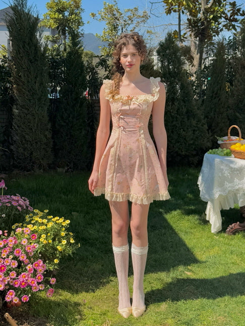A young woman in a vintage-inspired pink mini dress adorned with lace and ribbon details, standing gracefully in a garden. The dress features whimsical fairytale prints and a delicate lace hem, paired with lace knee-high socks for a romantic, cottagecore look.