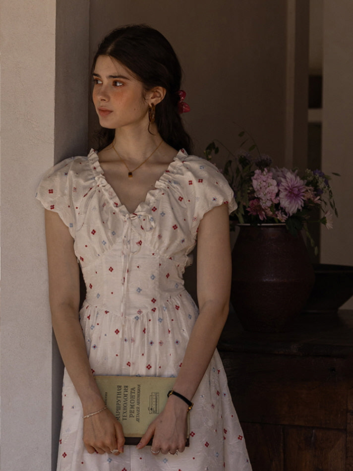 Close-up of a young woman in a cream floral print dress with a ruffled neckline and puff sleeves, holding a vintage book while leaning against a wall. A vase of flowers adds a romantic, vintage-inspired touch to the scene.
