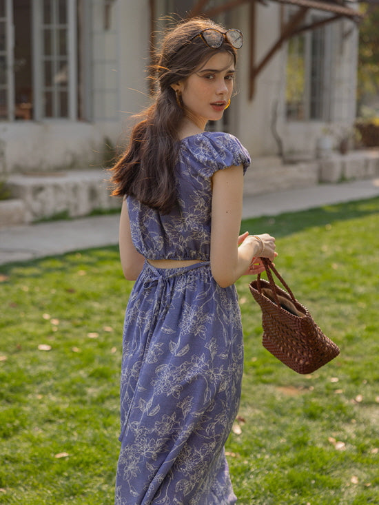 Back view of a woman in a blue floral cut-out dress, carrying a woven bag while strolling through a sunlit garden, capturing a dreamy, vintage-inspired vibe.