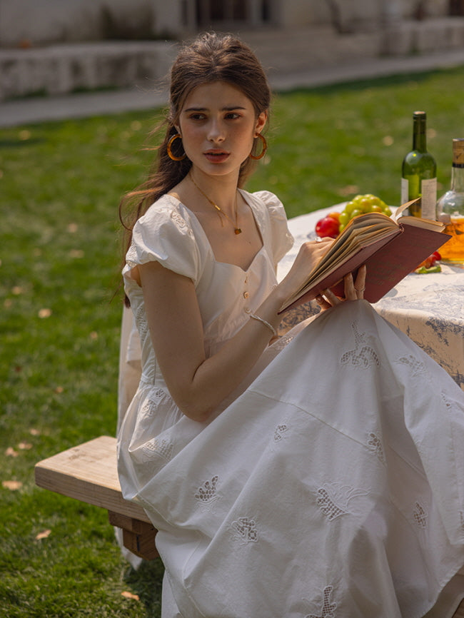 A woman in a vintage-inspired white embroidered dress with cap sleeves, sitting on a wooden bench while reading a book. The dress features delicate floral cutwork details and a romantic cottagecore aesthetic.
