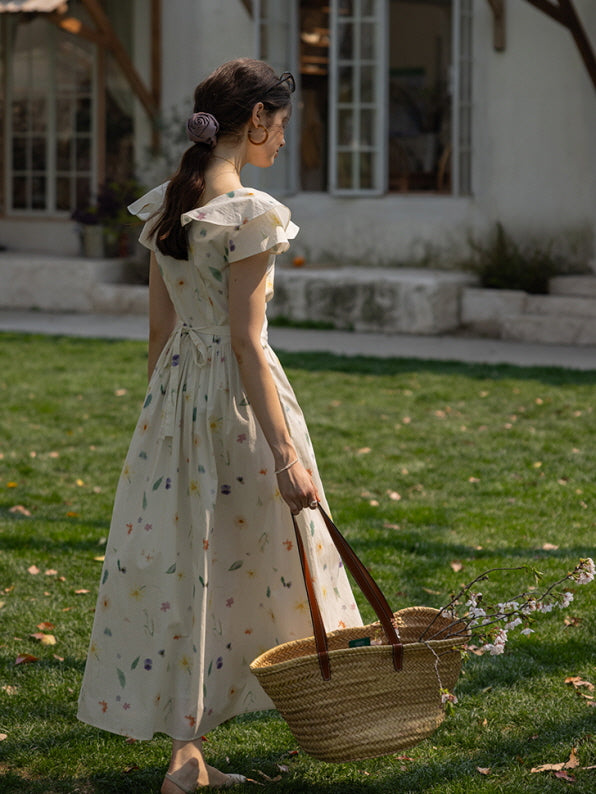 Rear view of a woman in a cottagecore-inspired floral dress with delicate ruffled sleeves, holding a woven basket filled with blooming branches while strolling through a garden.