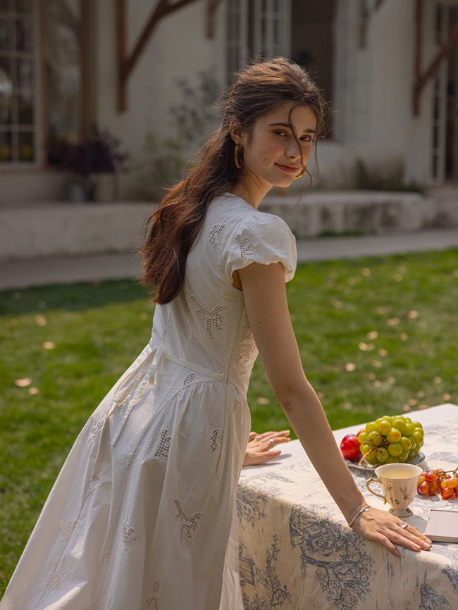 A woman in a white embroidered dress with cap sleeves, leaning over a picnic table adorned with fresh fruits and a vintage floral tablecloth.