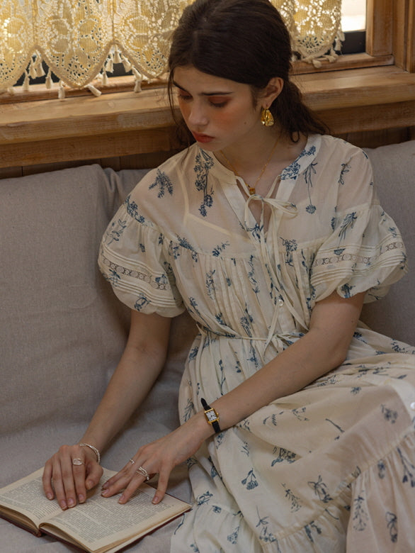 Young woman sitting on a cozy sofa in a cream floral print dress with blue botanical patterns and puff sleeves, reading a book while surrounded by lace curtains, evoking a soft, cottagecore vibe.