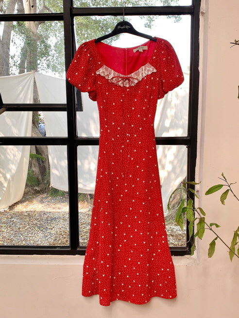 A red retro dress with lace-trimmed neckline and puff sleeves, shown hanging by a window with natural light, capturing its elegant silhouette and summery charm.