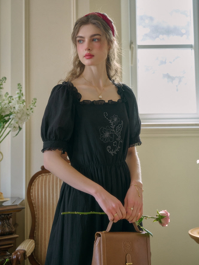 A young woman with wavy hair and a red headband holding a pink rose and a brown retro handbag, wearing a black vintage-inspired dress with floral embroidery, standing near a sunlit window.