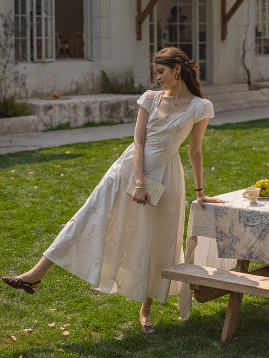 Woman playfully stretching her leg while holding a book, standing by a picnic table covered in a floral tablecloth, dressed in a white, vintage-style dress with floral embroidery.
