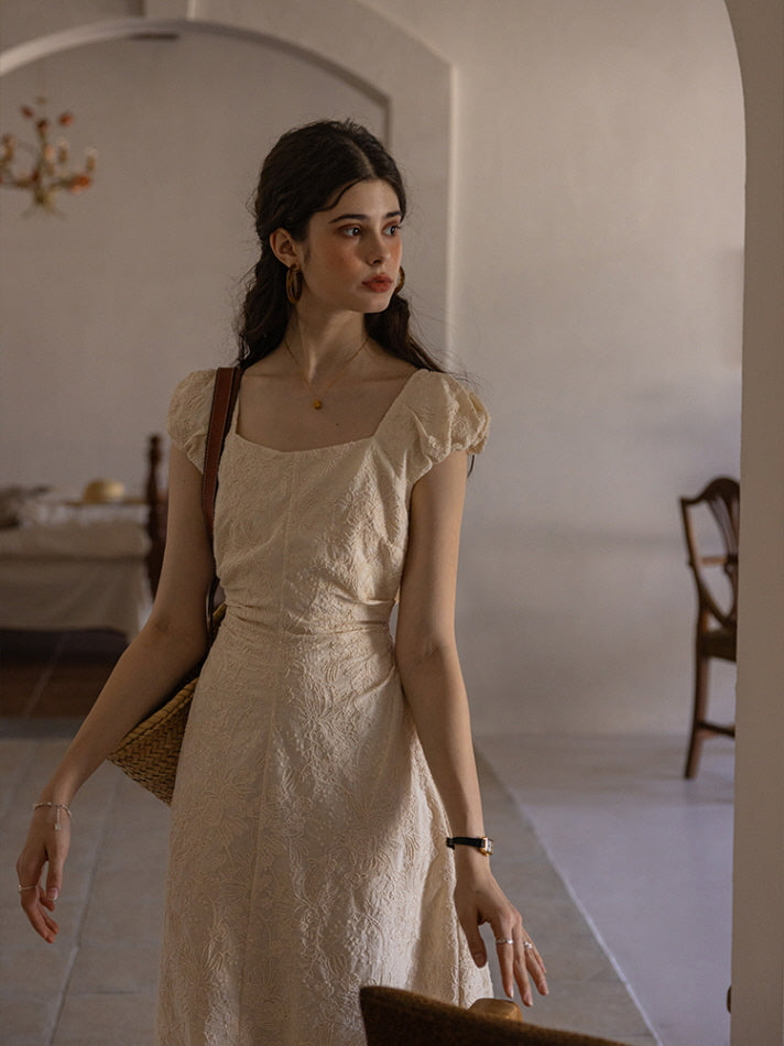 Woman wearing a cream-colored textured midi dress with puff sleeves and a square neckline, carrying a woven tote bag while walking through a vintage-inspired, softly lit interior space.