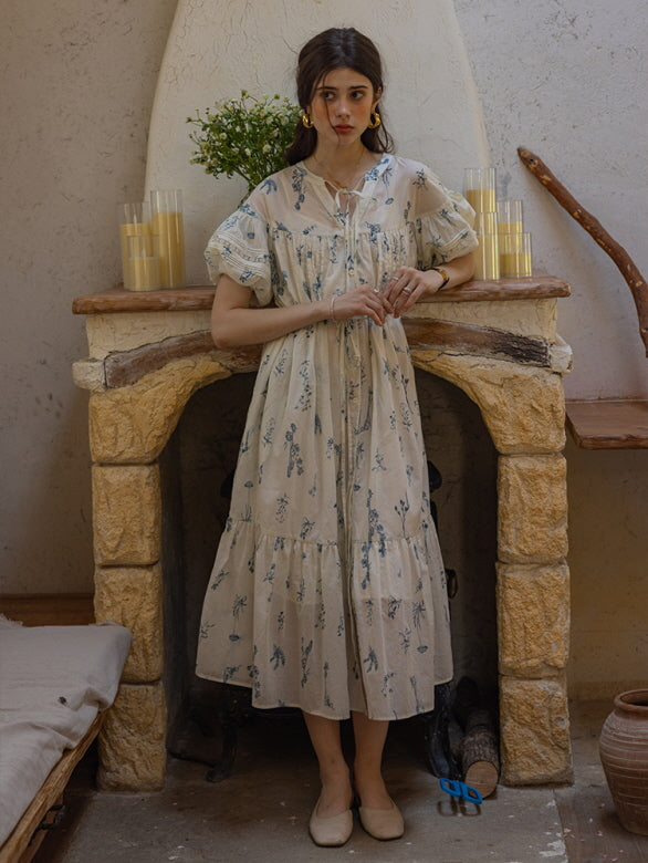 Young woman standing in front of a stone fireplace wearing a cream floral print dress with tiered skirt and puff sleeves, surrounded by candles and rustic decor. The setting exudes a romantic, countryside charm.