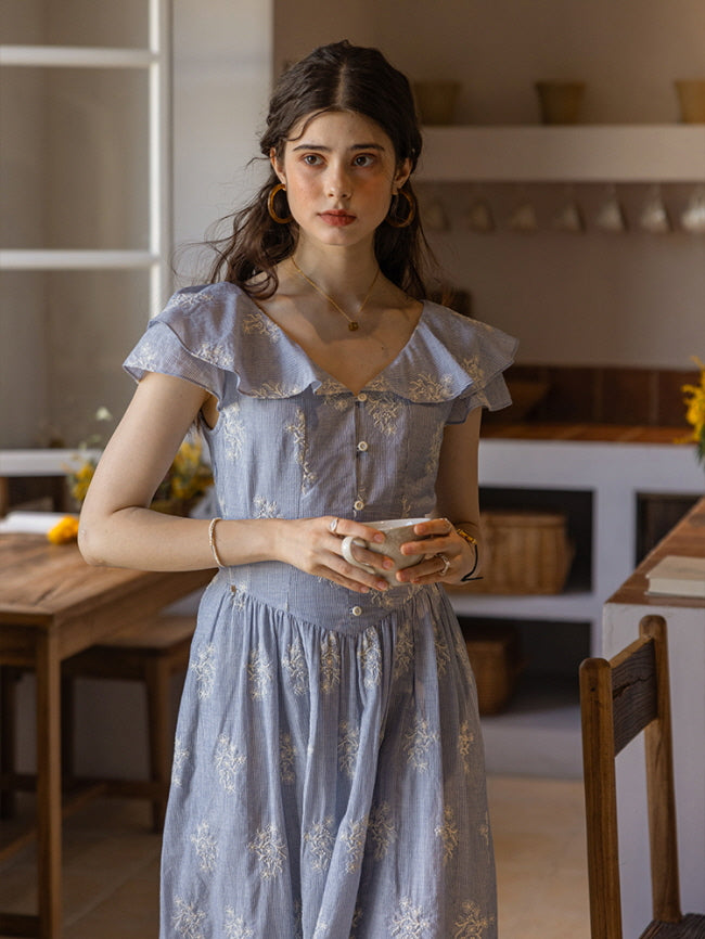 A young woman with soft, wavy hair gazes thoughtfully out the window, holding a ceramic teacup in her hand. She wears a light blue floral cotton dress with a ruffled collar and button-down front, evoking a vintage, cottagecore charm. An open book and fresh lemons rest on the wooden counter beneath her.