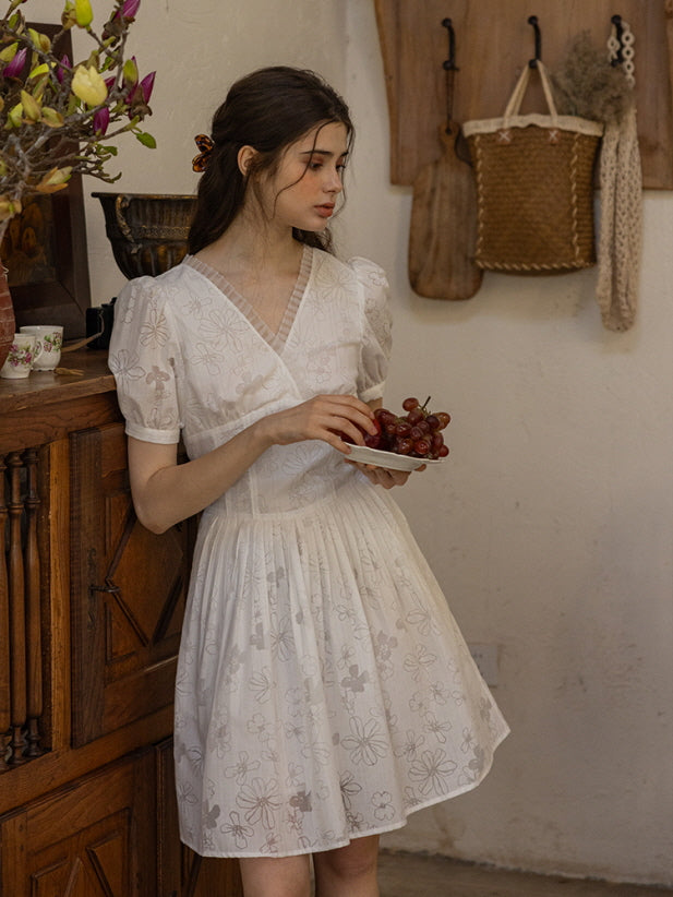 Young woman holding a plate of grapes while wearing a white floral mini dress with a wrap-style bodice and pleated skirt, exuding a vintage summer look in a cozy, cottage-style setting