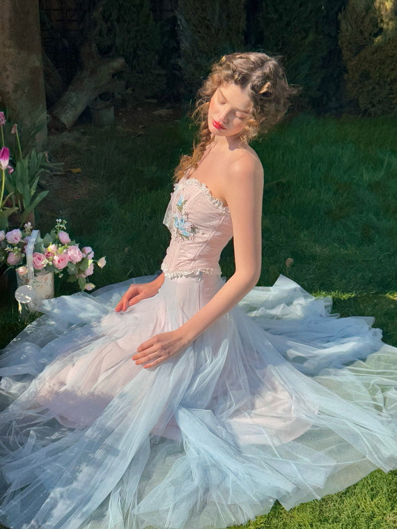 Elegant woman sitting on the grass in a pastel floral corset dress with blue tulle skirt, surrounded by soft sunlight and spring flowers – dreamy vintage aesthetic fashion