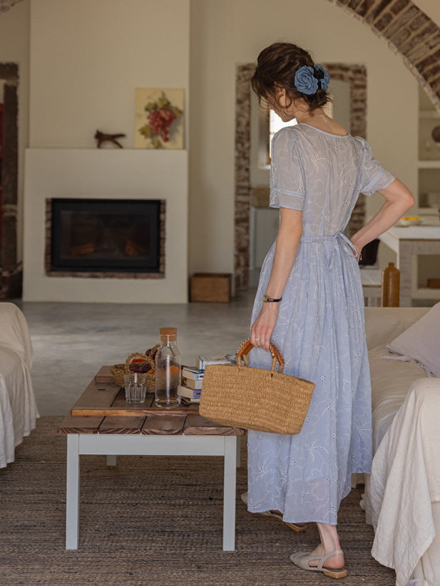 Back view of a woman wearing a blue eyelet lace dress tied at the waist, holding a woven basket bag in a cozy cottage-style room. Romantic, vintage-inspired look.