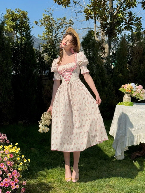 A young woman in a vintage-inspired floral cotton dress poses gracefully in a blooming spring garden, bathed in sunlight. Her romantic outfit with puff sleeves and a pink ribbon corset detail reflects a fairytale aesthetic, captured in a soft countryside setting.