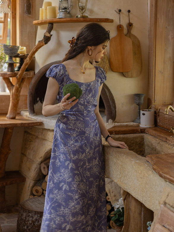 Woman in a blue floral dress holding a moss ball, standing by a rustic stone sink in a vintage-inspired cottage kitchen, exuding a countryside aesthetic.