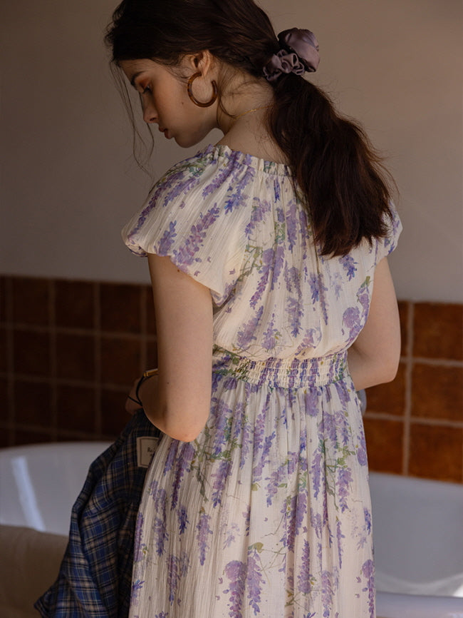 Back view of a lavender floral dress with soft ruffled sleeves and a cinched waist, evoking a romantic cottagecore style against a muted indoor backdrop.