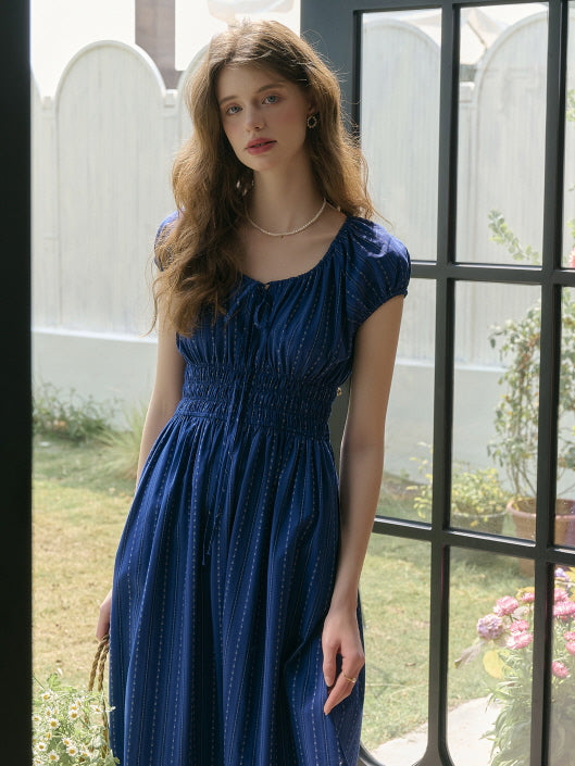 Model in a navy blue cap-sleeve dress with gathered bodice and flowy skirt, standing by a window with a peaceful garden in the background. Elegant countryside mood with vintage flair.