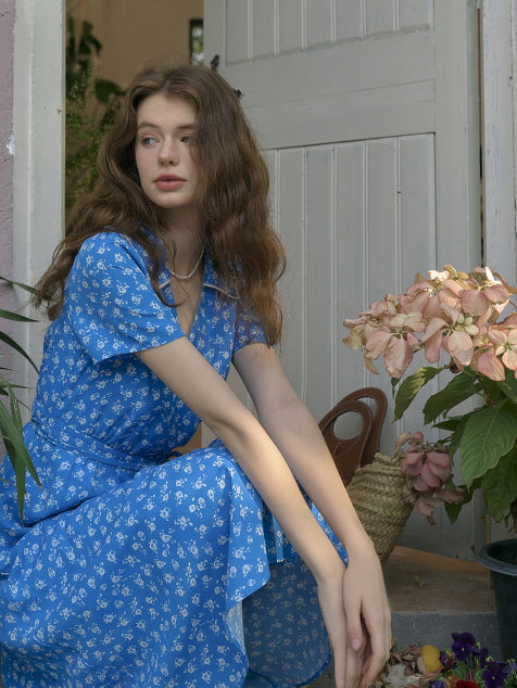 Young woman in a blue floral vintage-style dress sitting gracefully by a door, surrounded by soft pink hydrangeas and greenery. Dreamy cottagecore summer look with natural lighting.