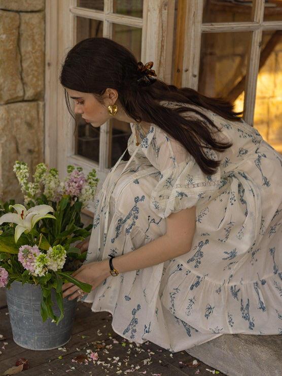 Woman in a cream floral print dress with blue botanical patterns, kneeling down to arrange flowers in a rustic metal bucket on a wooden porch. The scene evokes a vintage, cottagecore aesthetic.