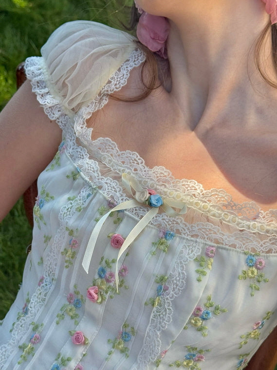 Close-up of a floral lace corset top with sheer puff sleeves, adorned with pastel floral embroidery and delicate ribbon bow detail at the neckline, capturing a soft, romantic aesthetic.