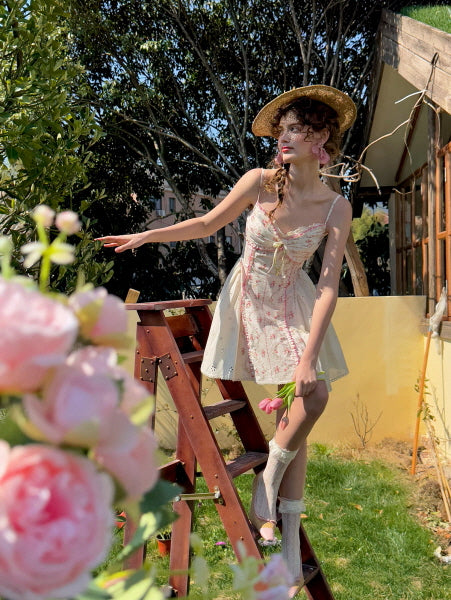 Coquette floral mini dress with lace and ribbon details, styled with a straw hat and lace socks. Vintage-inspired summer aesthetic, capturing playful, romantic vibes amidst a garden backdrop.