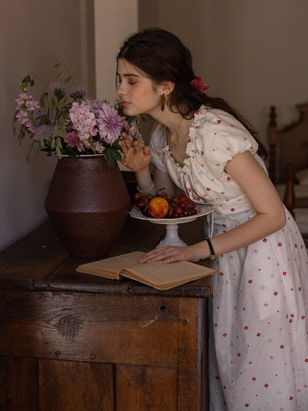 Woman in a cream floral print dress with ruffled neckline, leaning forward to smell a bouquet of flowers in a rustic vase. An open book and a plate of fresh fruits add a charming, vintage-inspired cottagecore aesthetic.