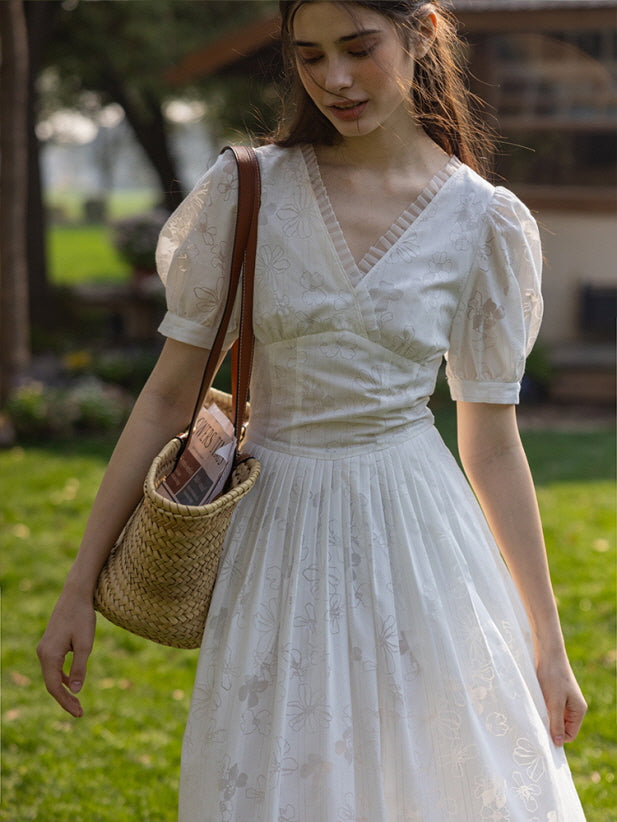 Close-up of a woman wearing a white floral embroidered dress with puff sleeves, holding a woven basket bag with a magazine inside.