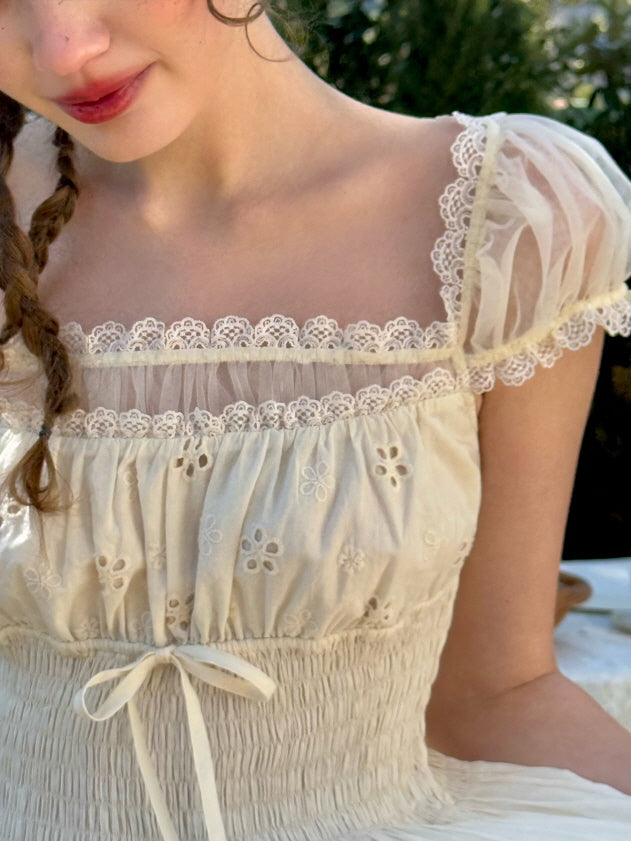 Close-up of a puff-sleeve ivory dress featuring delicate lace trim along the neckline, sheer tulle details, and daisy eyelet embroidery with a gathered waist and ribbon bow.