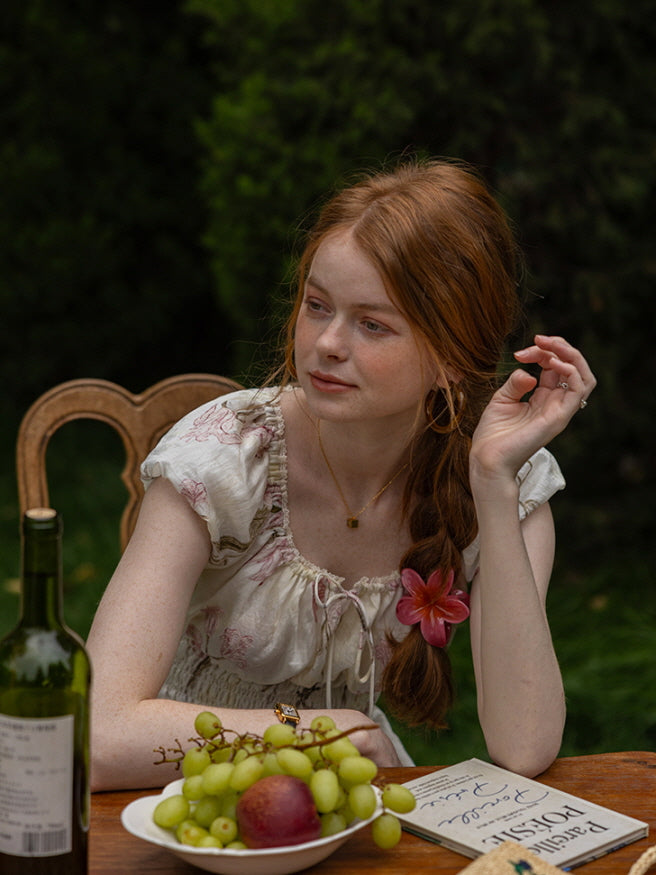 A woman seated at a wooden table adorned with grapes and wine, wearing a floral cottagecore dress with a ruffled neckline and floral hair accessory. Dreamy, vintage-inspired look.