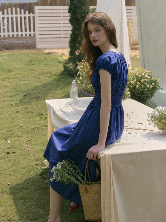 Woman sitting gracefully on an outdoor table draped in ivory fabric, wearing a navy shirred-waist dress and holding a straw bag with fresh flowers. Serene fairytale garden scene with soft sunlight.