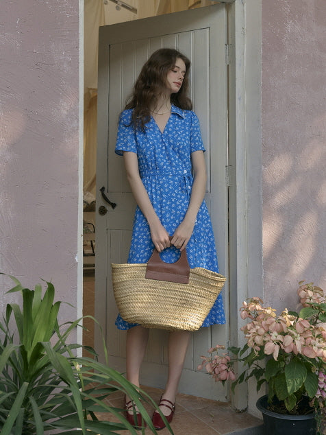 Woman standing in front of a pastel pink wall and wooden door, wearing a blue floral dress and holding a straw tote bag. Romantic vintage outfit styled with soft shadows and garden flowers.