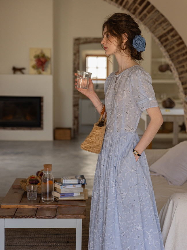 Side view of a woman holding a glass of water, wearing a blue eyelet lace dress with puff sleeves and a woven basket bag. Cozy cottagecore interior.