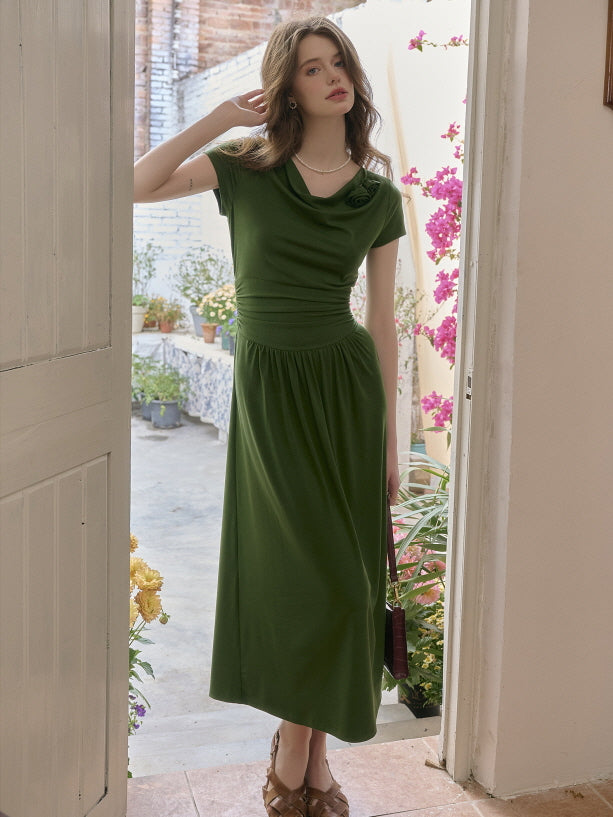 Woman in an elegant forest green draped neckline dress with a cinched waist, standing at a sunlit doorway while touching her hair. Romantic garden-inspired look with a soft, vintage charm.