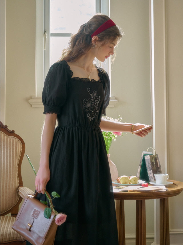 A young woman in a black retro vintage dress with puff sleeves and floral embroidery, standing by a sunlit window while holding a brown leather handbag and a pink rose.