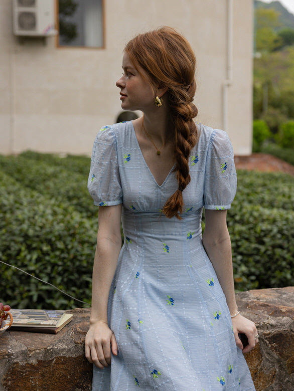 Side profile of a woman seated on a stone wall, wearing a light blue floral midi dress with puff sleeves. Her long braided hair and delicate accessories evoke a vintage summer vibe.