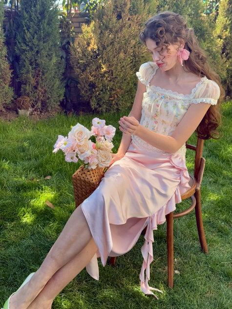 A model seated in a garden chair, wearing a floral lace corset top with sheer puff sleeves and pastel floral embroidery, styled with a pink ruched satin skirt adorned with ribbon ties, holding a woven basket filled with delicate pink and cream roses, creating a soft, romantic aesthetic.