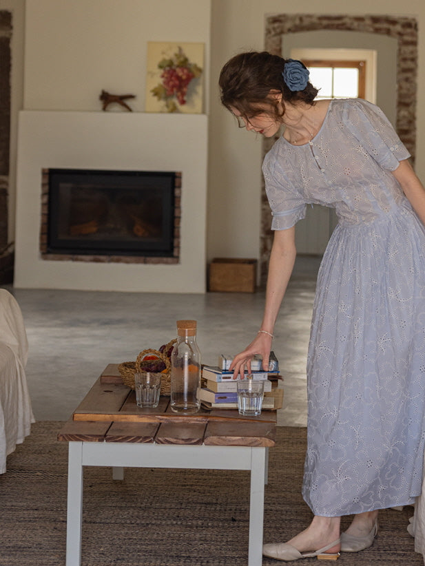 Woman leaning over a wooden table, arranging books and glasses while wearing a blue eyelet lace dress with puff sleeves. Romantic, vintage-inspired look.