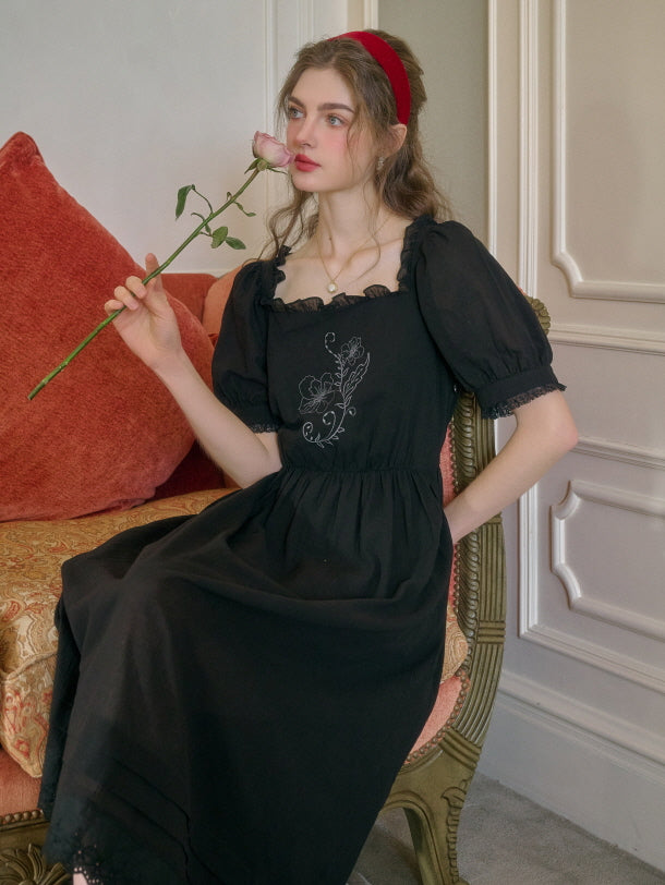 A young woman sitting gracefully on a vintage armchair, wearing a black retro-style dress with floral embroidery, holding a pink rose and gazing thoughtfully to the side.